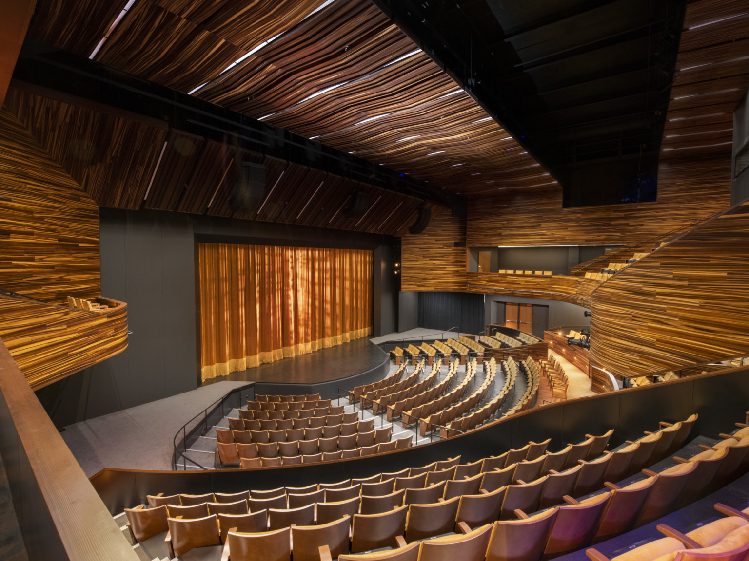 View from back of theater to stage with yellow brown velvet curtain. Panels of textural wood make up the ceiling