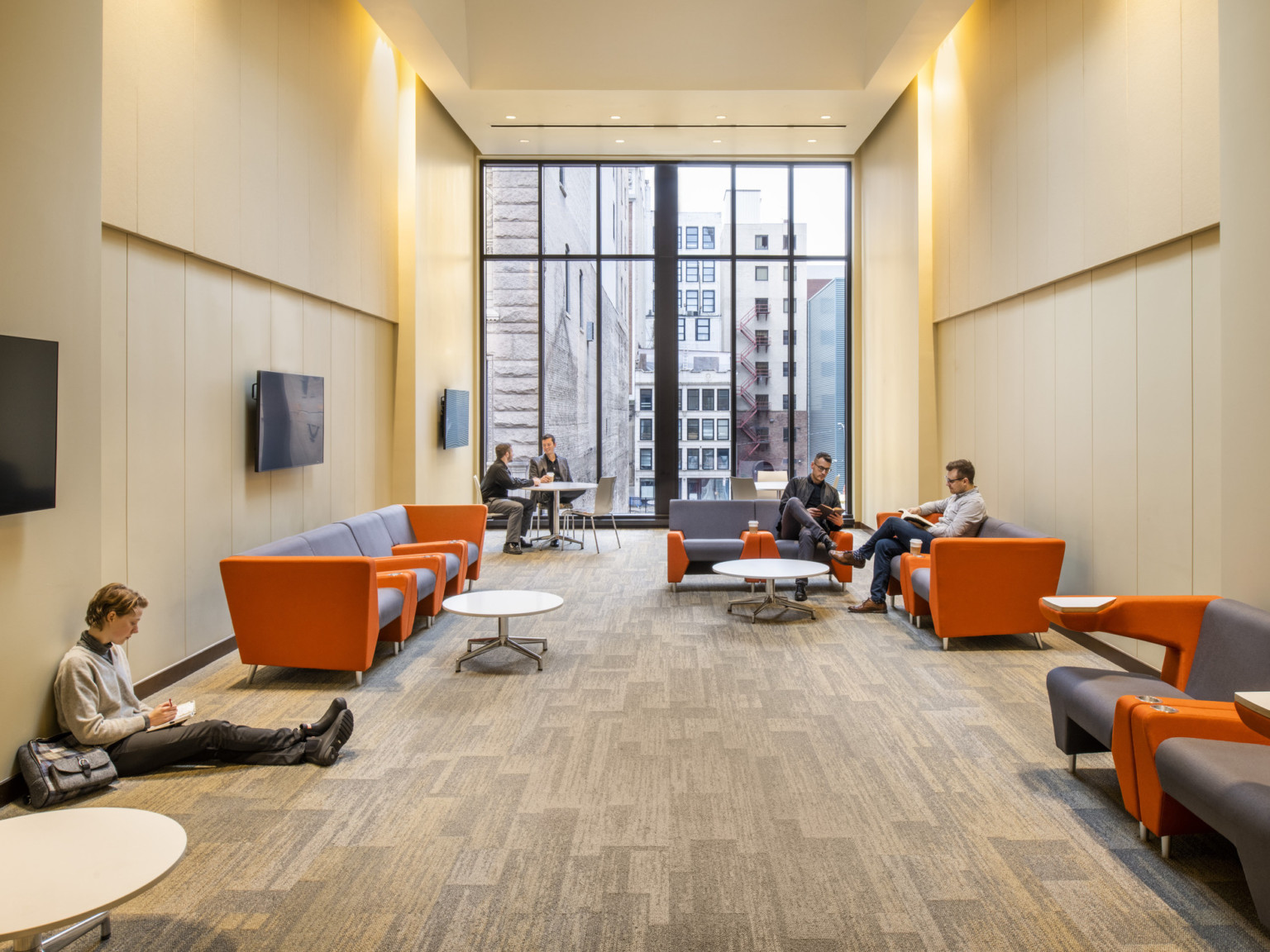 Carpeted hallway with floor to ceiling window, multiple couches with coffee tables, and screens along the wall