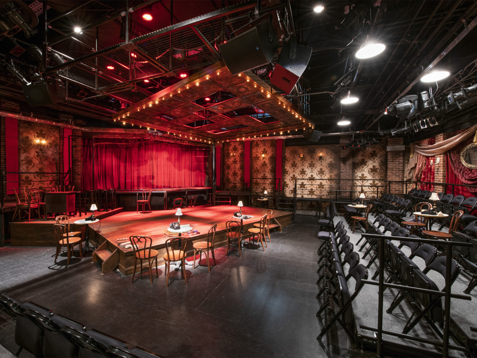 A black box theater with exposed fly system and light fixtures, the stage is set with tables and chairs