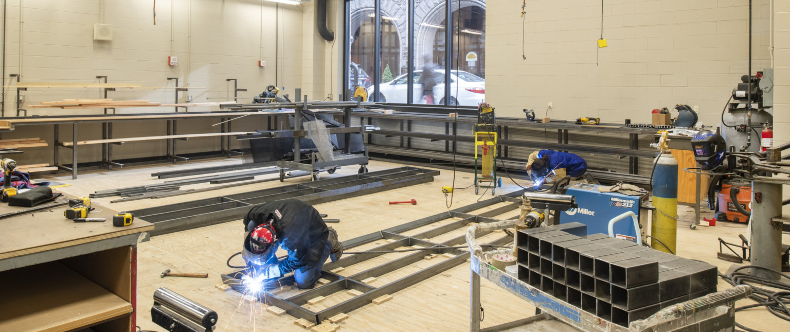 Workspace with welding equipment and tools, metal is being worked. A large window to the back looks out on street level