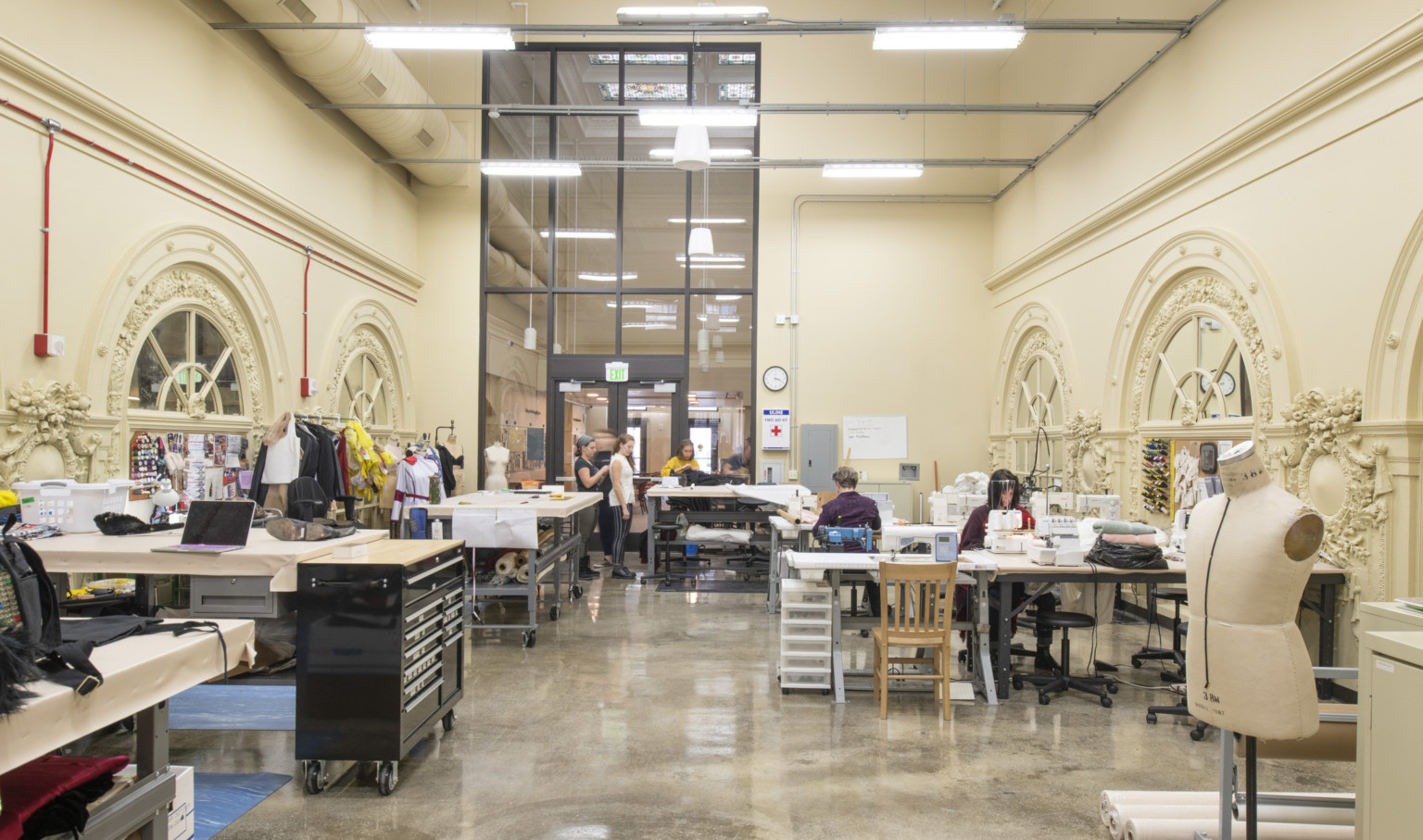 Double height long room with sewing machines and arched mirrors with ornate molding details on either side of work tables Double height long room with sewing machines and arched mirrors with ornate molding details on either side of work tables