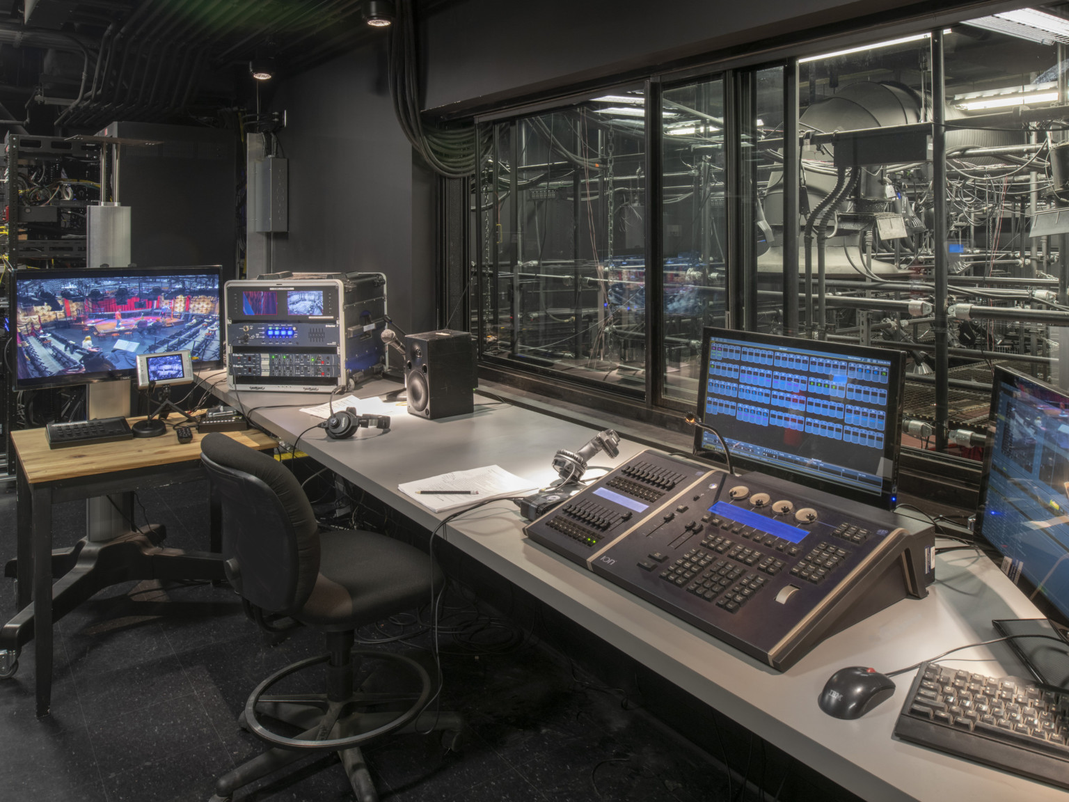 A control panel sits on top of a long table with multiple screens in front of a window looking into a room with grey pipes