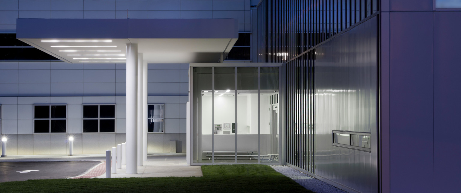 Side view of entry with glass vestibule with benches. Overhang above entry to driveway. Grey wall behind with square windows