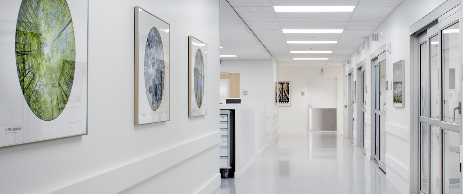 White hallway with doors to right. Room to left with medical cart under counter. White door with metal bottom at end of hall