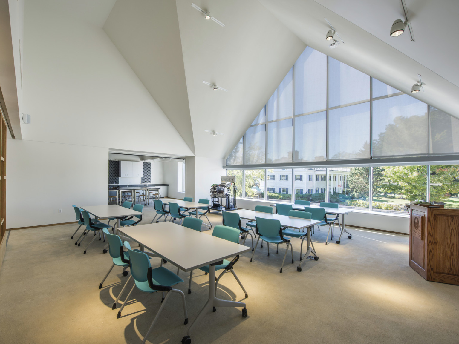 White double height room with vaulted ceilings and large window coming to a point. Wood accent wall left. Tables and chairs