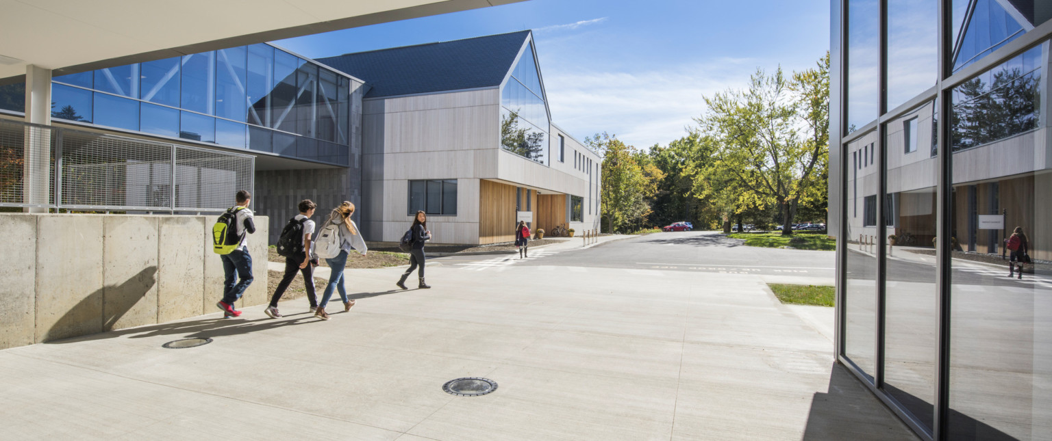 Exterior covered walkway between 2 buildings. Floor to ceiling windows, right. Left, an elevated hallway to stone building