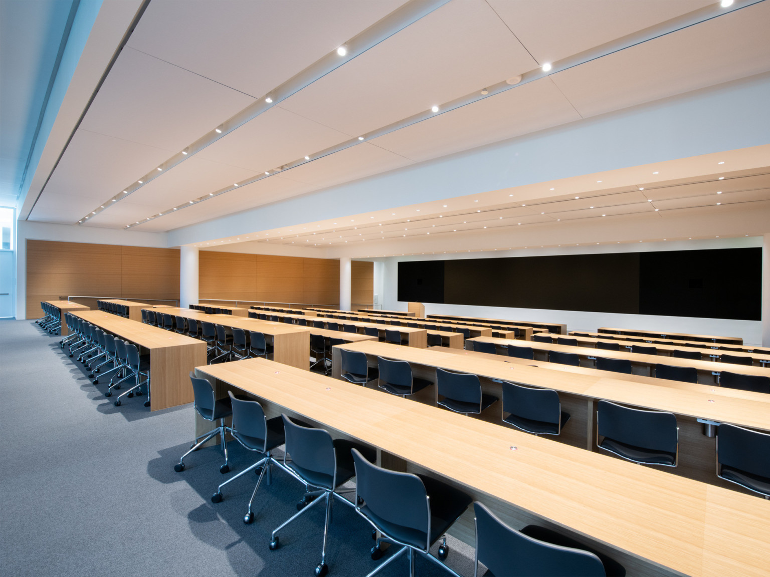 White room with wood accent on left wall. Long tables with chairs face podium and wide screen on front wall