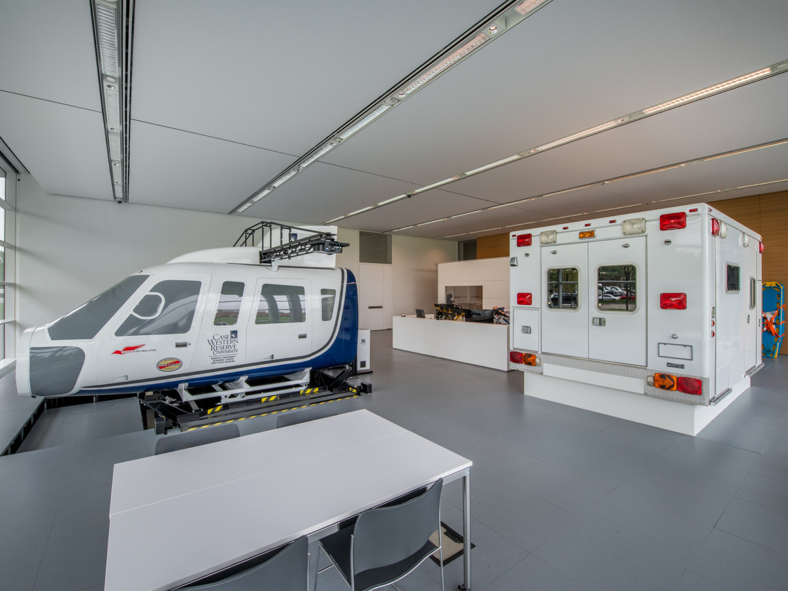 White room with grey floor. Cockpit and passenger area of dismantled helicopter left, ambulance cabin right, a table between