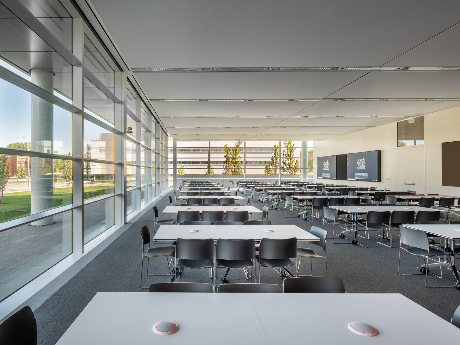 White room with 2 walls of floor to ceiling windows and drop ceiling panels. Tables and chairs line both sides of room