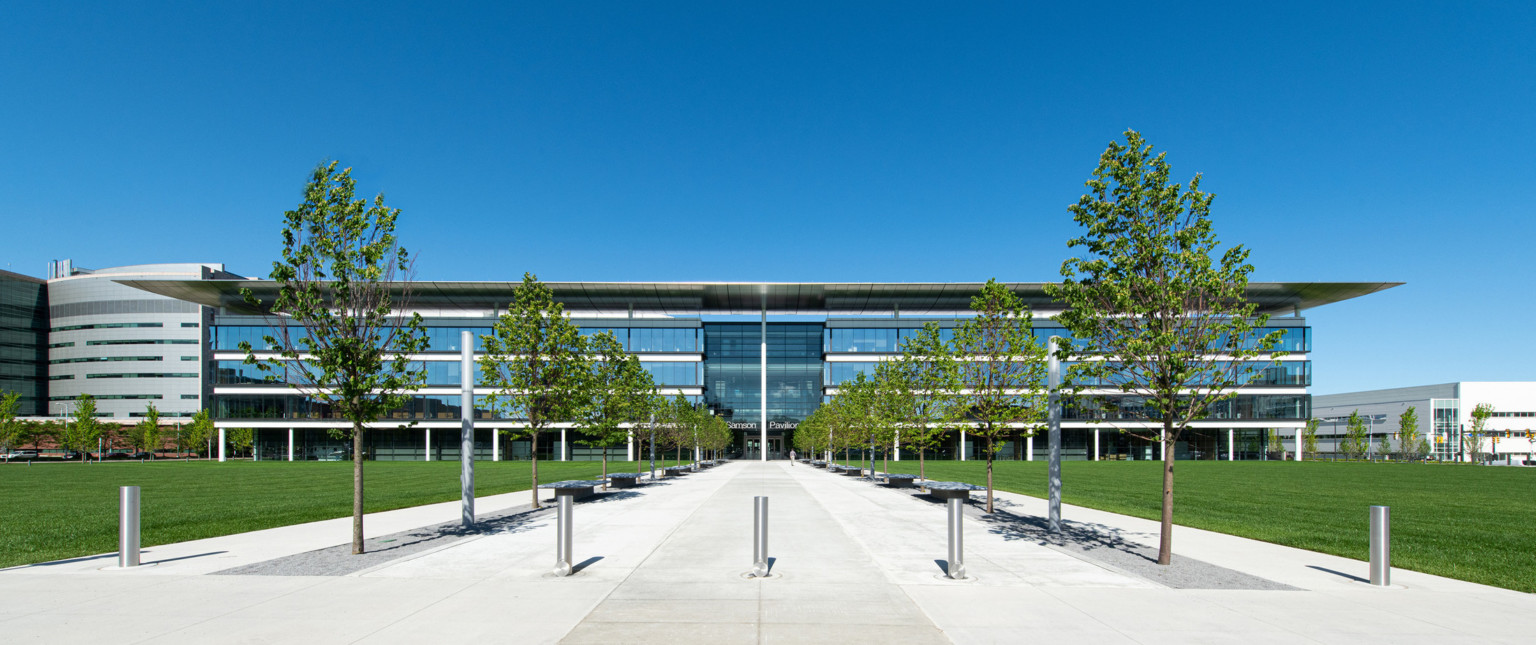 Building front with 4 stories of floor to ceiling windows and recessed double height window entry. Tree lined walkway to door