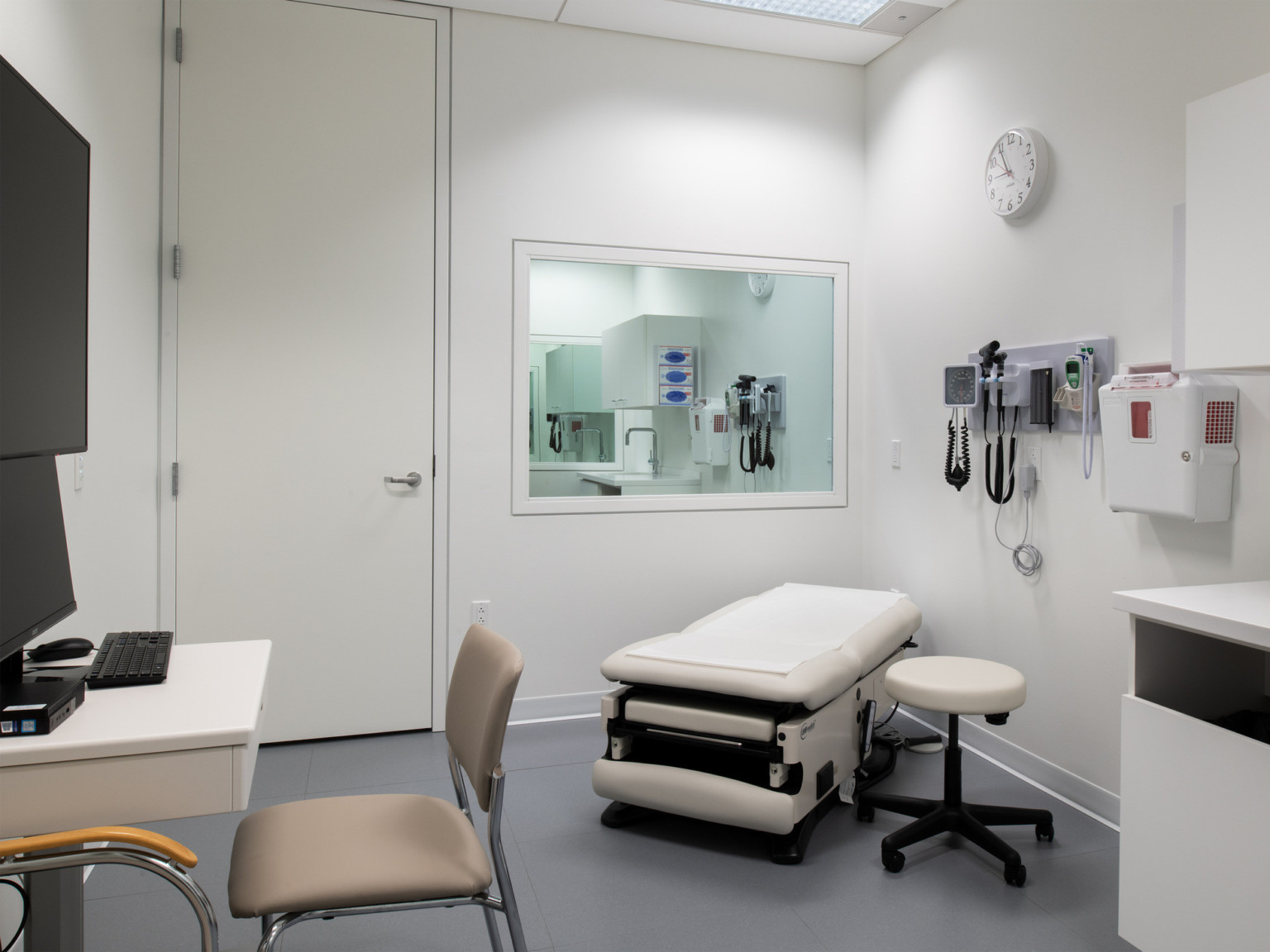 Exam room with medical bed and matching stool. Equipment hangs on wall above, mirror on left wall reflects faucet. Desk left