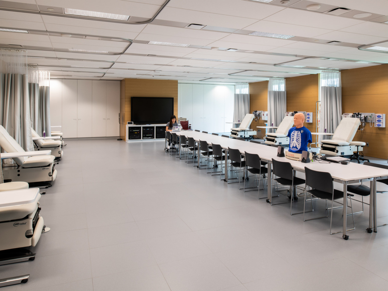 Reclined hospital beds line white room with wood accents and face long table with CPR dummy. Large screen on back wall