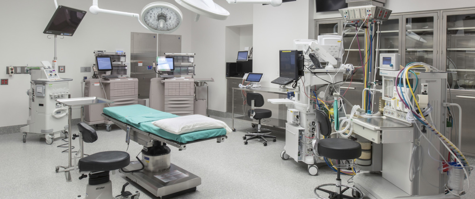 Operating room with table center, circular light above. Machinery stands along side the room, along with metal shelves