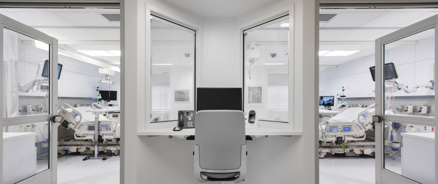 Desk with view into 2 hospital rooms, each with beds and screens. The white desk fits into a triangular space between doors