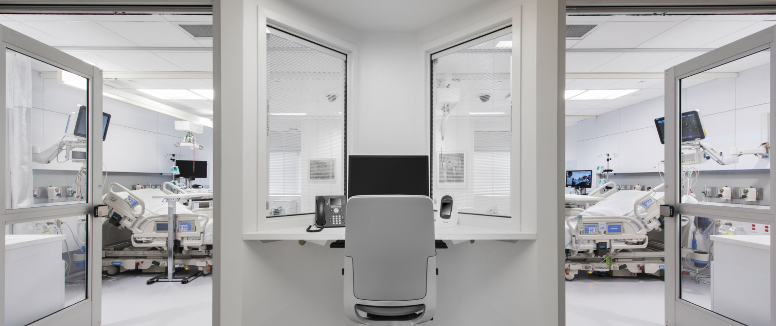 Desk with view into 2 hospital rooms, each with beds and screens. The white desk fits into a triangular space between doors Desk with view into 2 hospital rooms, each with beds and screens. The white desk fits into a triangular space between doors