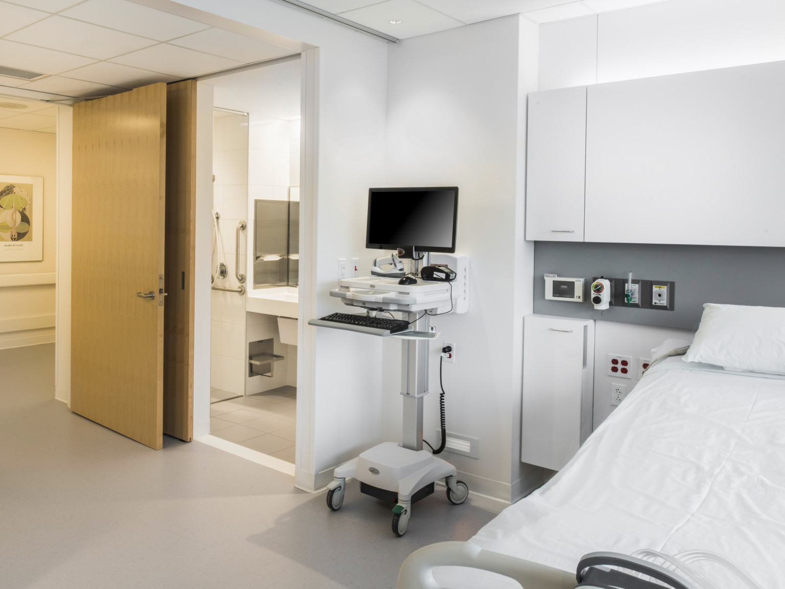 Inside a hospital with door in left corner. Private restroom is right of the door, with flexible workstation between the bed