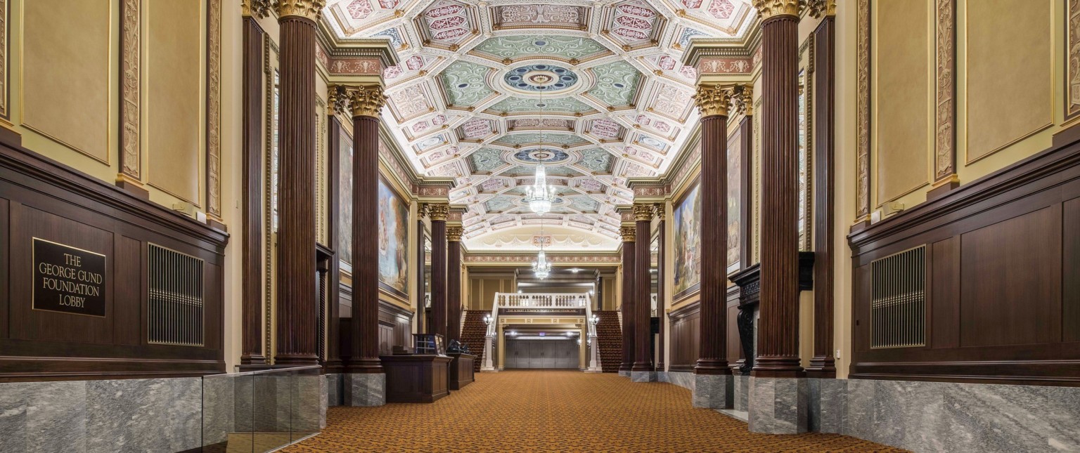 Grand staircase carpeted beneath ornate multicolored coffered ceiling with chandelier framed by ribbed pillars