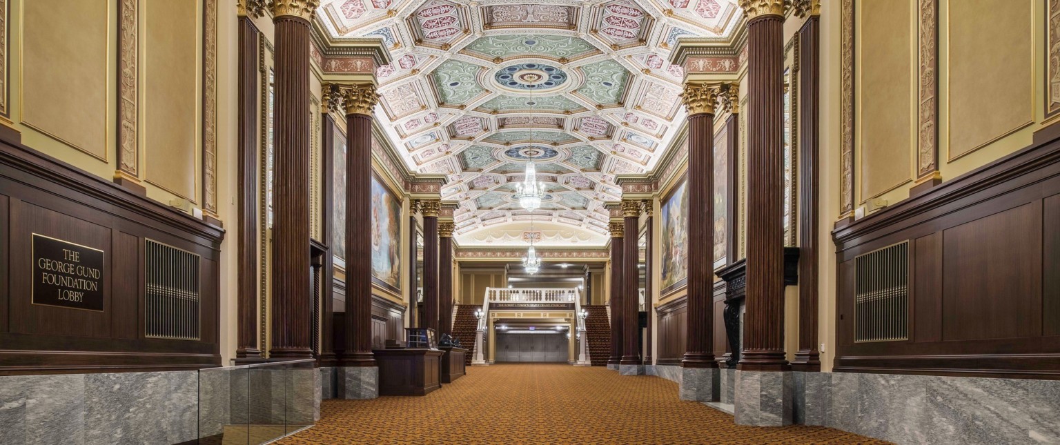 Grand staircase carpeted beneath ornate multicolored coffered ceiling with chandelier framed by ribbed pillars