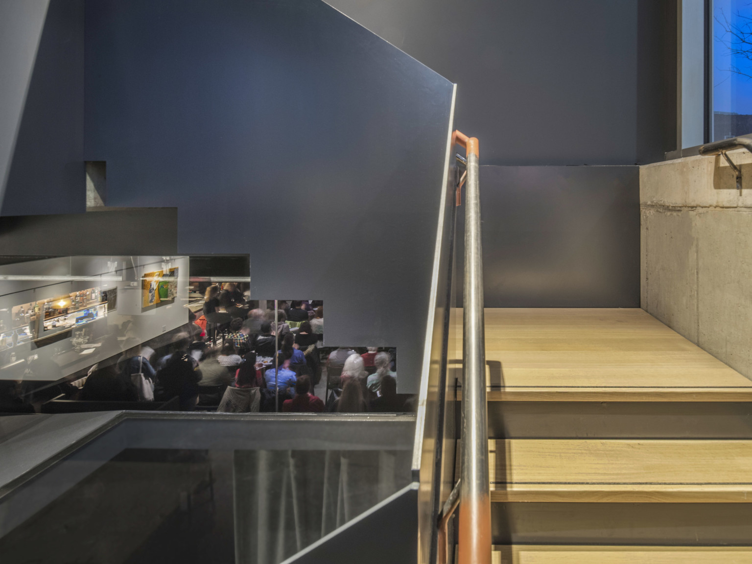 Wood stairs in stairwell with grey walls featuring stepped cutouts underneath, opening the stairwell to performance space