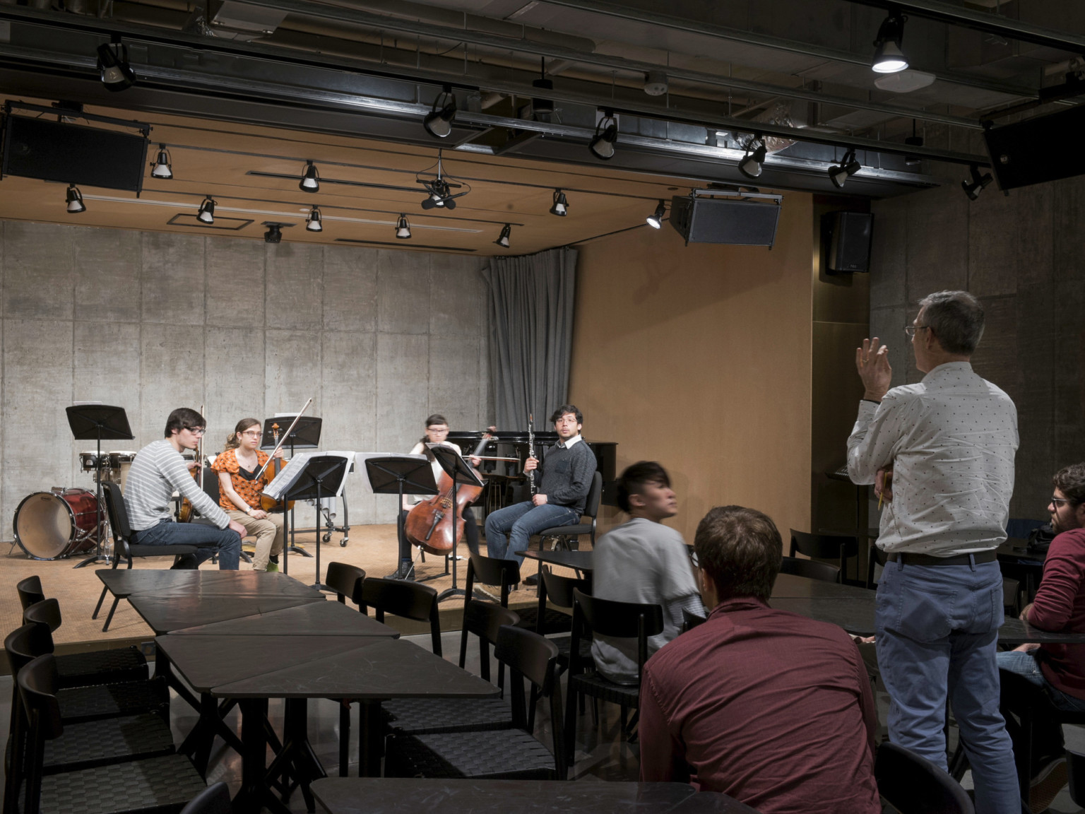 Teacher speaking to musicians on wooden box stage with grey stone back wall. Black light fixtures come from stage ceiling