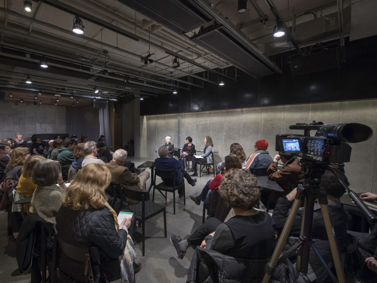 Audience and camera sit facing 3 individuals seated at front of concrete room with dark grey ceiling and exposed structuring