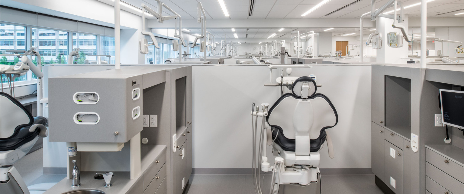 Dental chair with cleaning equipment in a grey cubicle. The tops of other cubicles and chairs can be seen. Large windows left