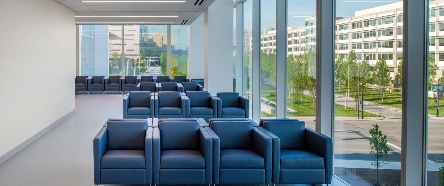 Seating area with blue arm chairs in white room with two walls of full length windows. Recessed lines of light from above