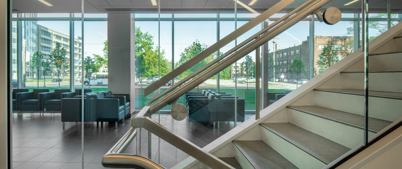 Room viewed through glass stair railing. Arm chair seating area, dark colored tiles, white walls, and floor to ceiling windows