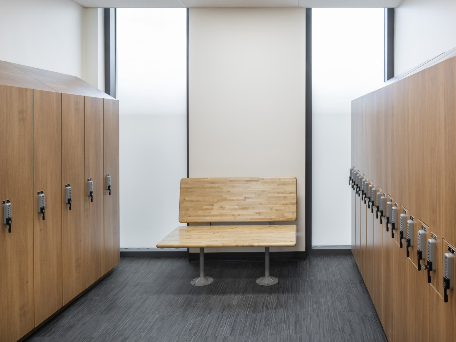 Room with wood lockers on both sides and wood bench center against white wall with translucent white windows on either side