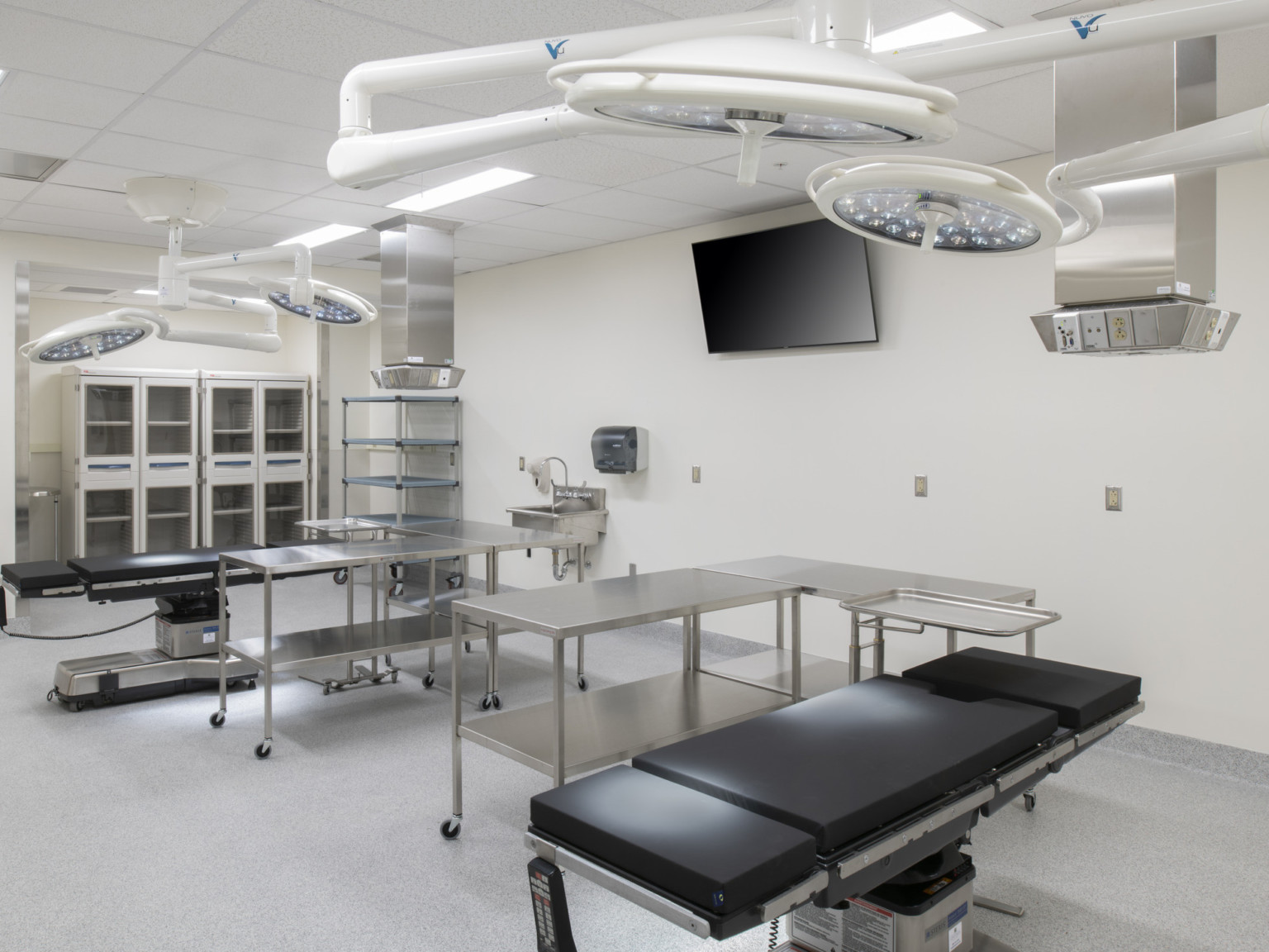 2 black hospital beds circular lamps above, metal tables next to each. White walls with metal cupboards in left corner