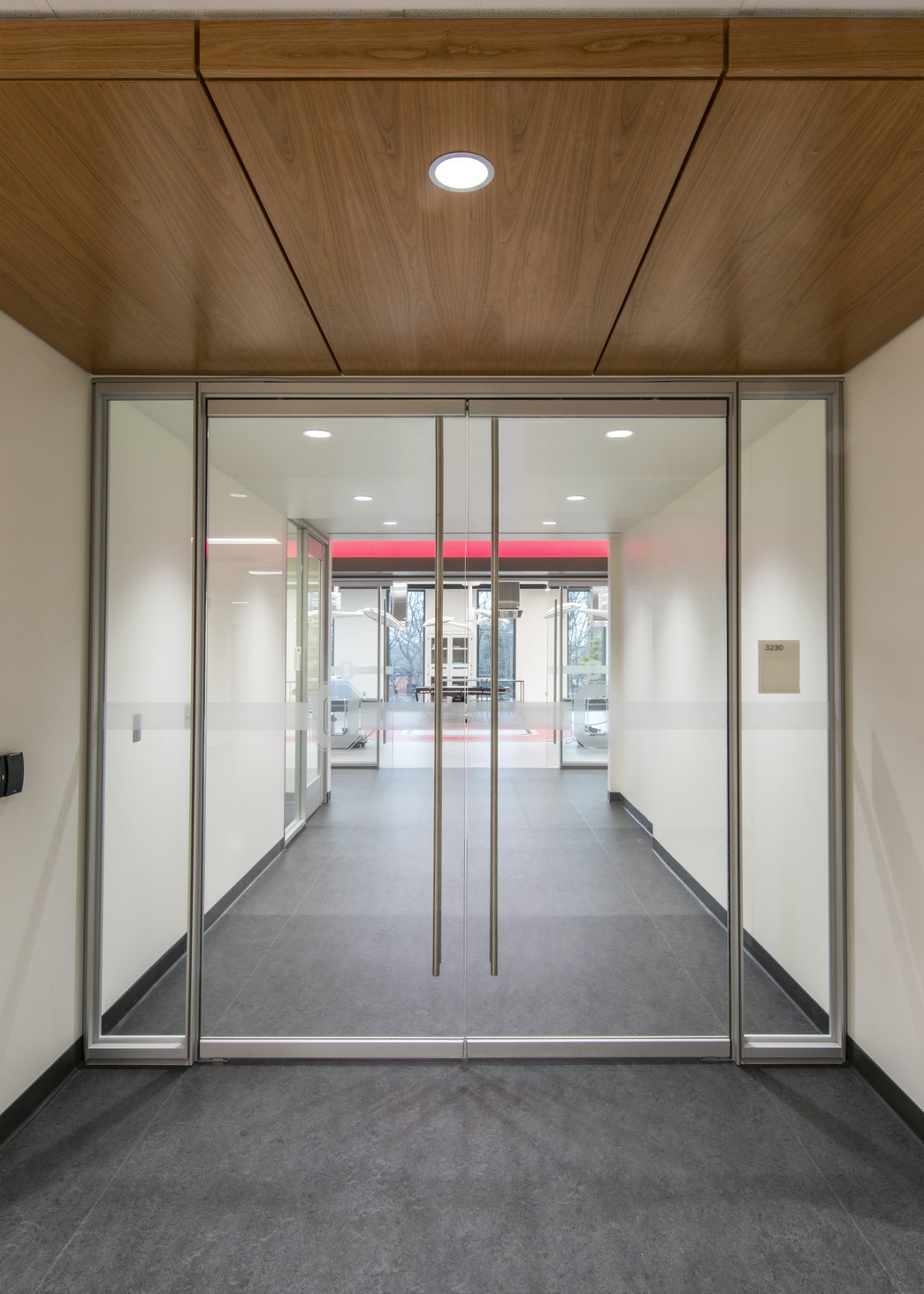 Grey carpeted hallway with glass double doors. Wood ceiling detail in front of doors with white ceiling beyond