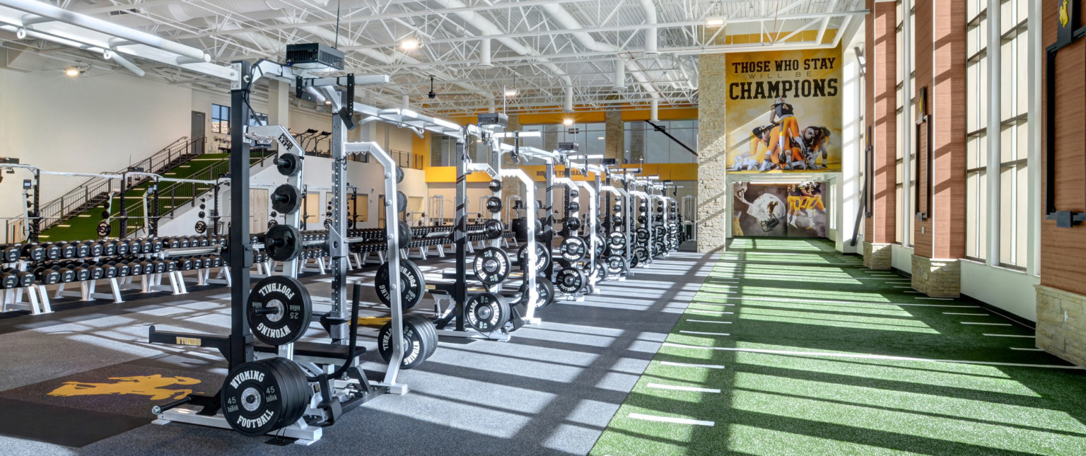 Turf floor with yard marks along double height windows. Ahead, a mural reading Those Who Stay Will Be Champions. Weights left