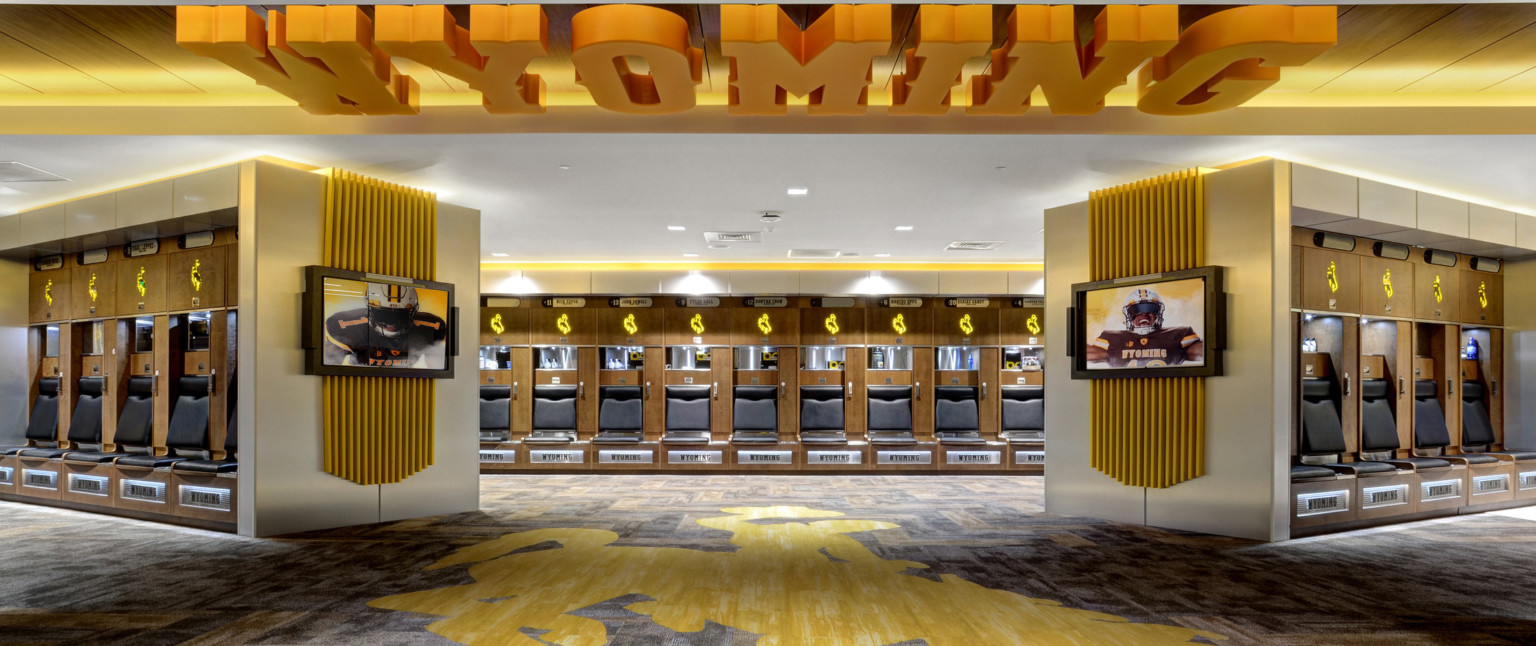 Locker room with yellow accents. Mascot illuminated above wood lockers. Yellow Wyoming sign on ceiling and mascot on carpet