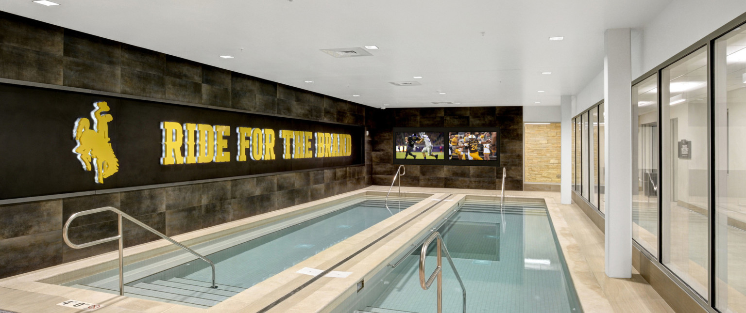 2 recovery pools in stone room with glass wall, right. Backlight logo, left, with Ride for the Brand phrase. Screens front