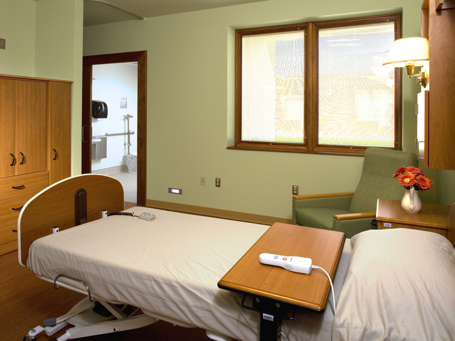 Light green room with bed and wood portable tray table. Green armchair right between sconce and window. Cabinet at bed foot.