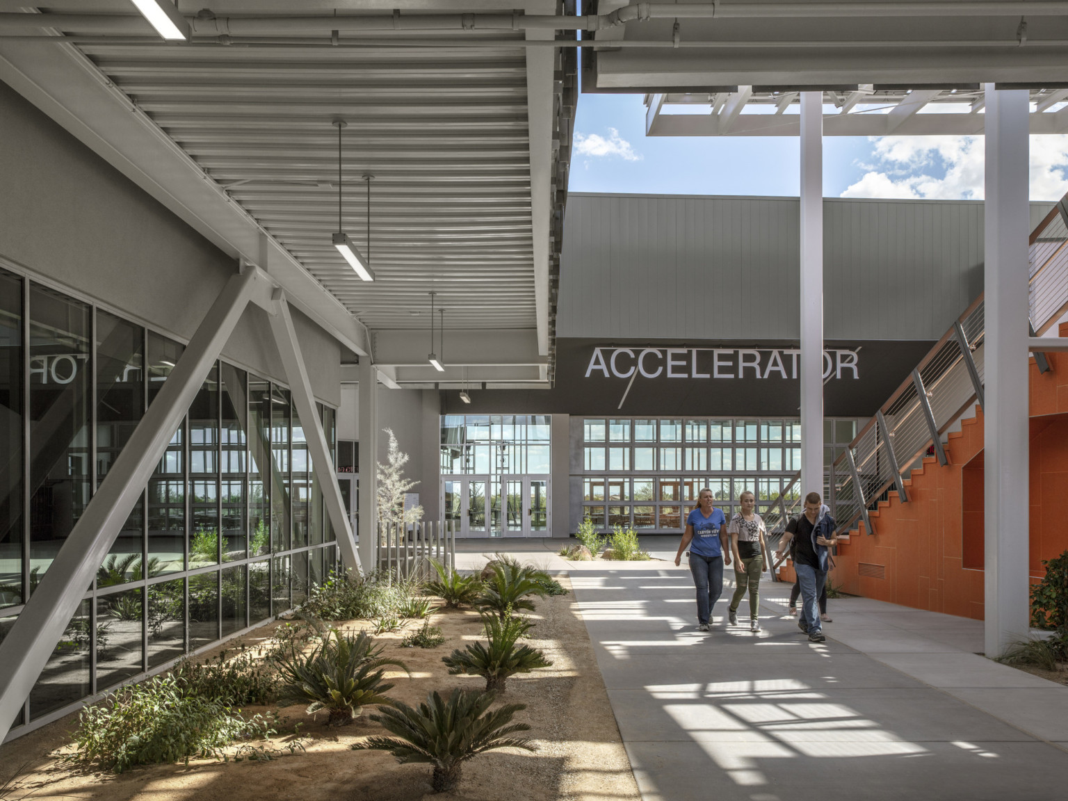 Breezeway to the left of the orange stairs. Behind is a building with a dark roof and parallel walls of double height windows