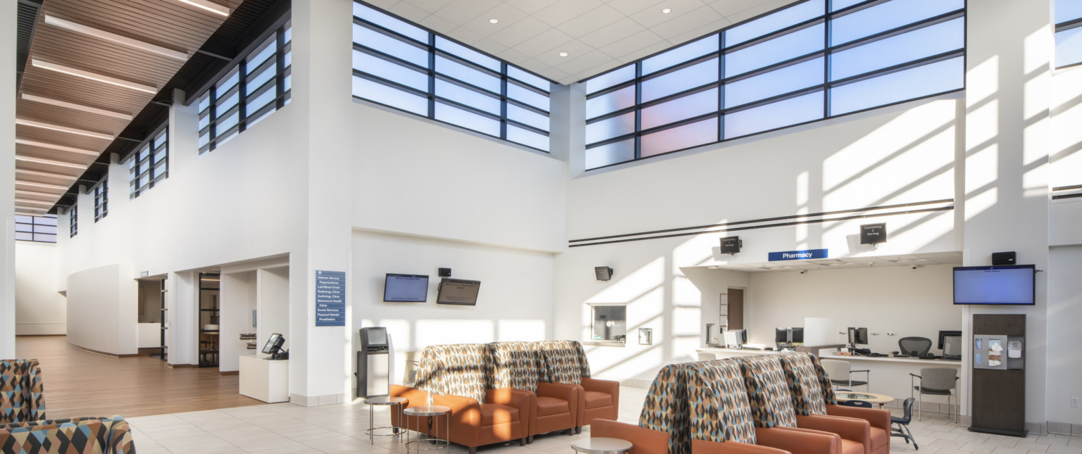 White room with double height ceiling and band of translucent windows around top. Armchair waiting area in front of pharmacy