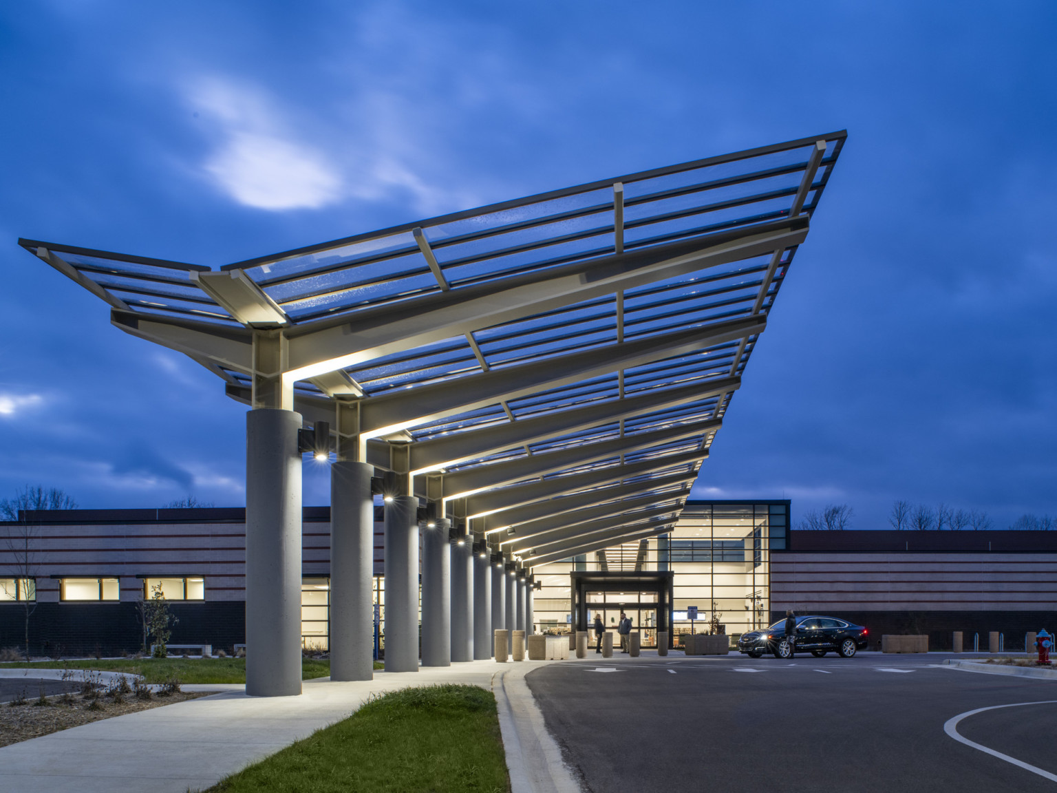 Entry viewed from driveway below angled transparent canopy with metal grid supported by grey pillars with large trusses