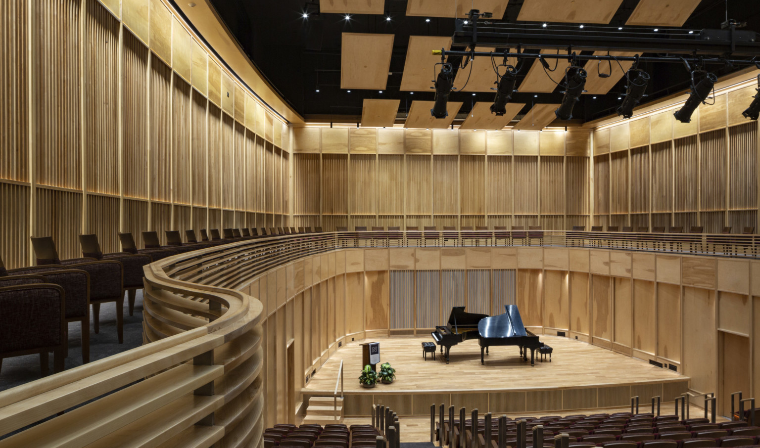 View looking down to performing stage with grand piano in a theater with all wood paneling and curved railing, with sophisticated sound customization panels at UNR Fine Arts and Music addition
