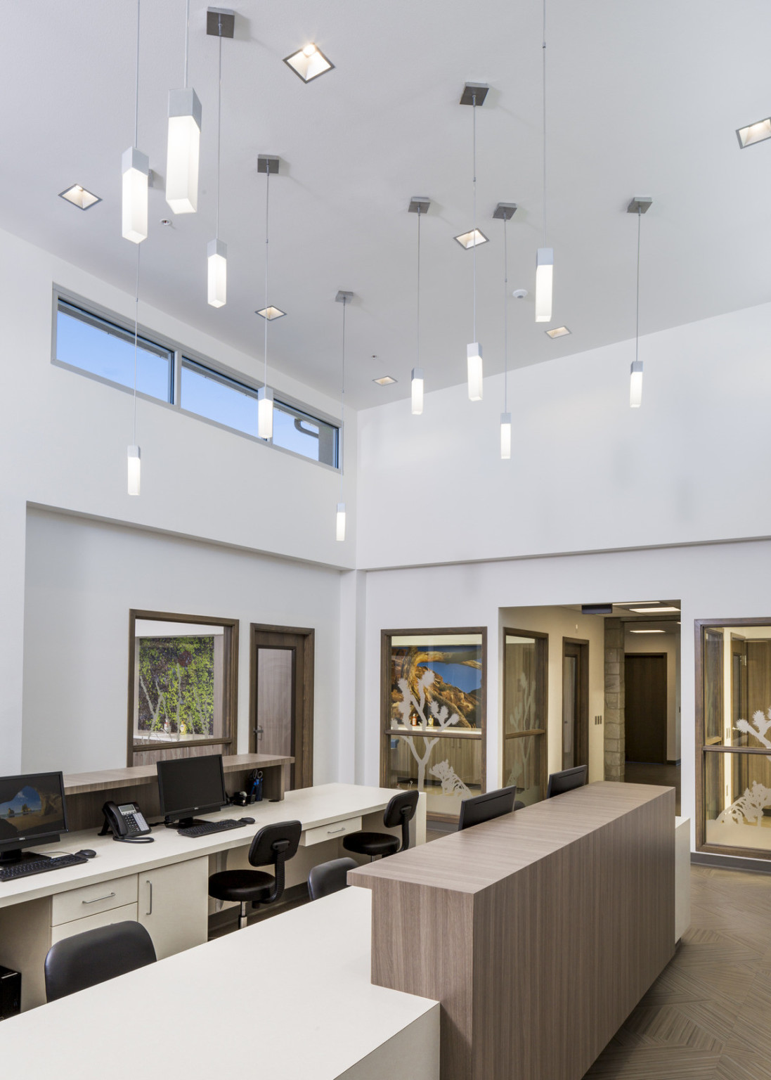 White room with desks and chairs under rectangular prism pendant lights with silver base and anchors. Interior windows behind