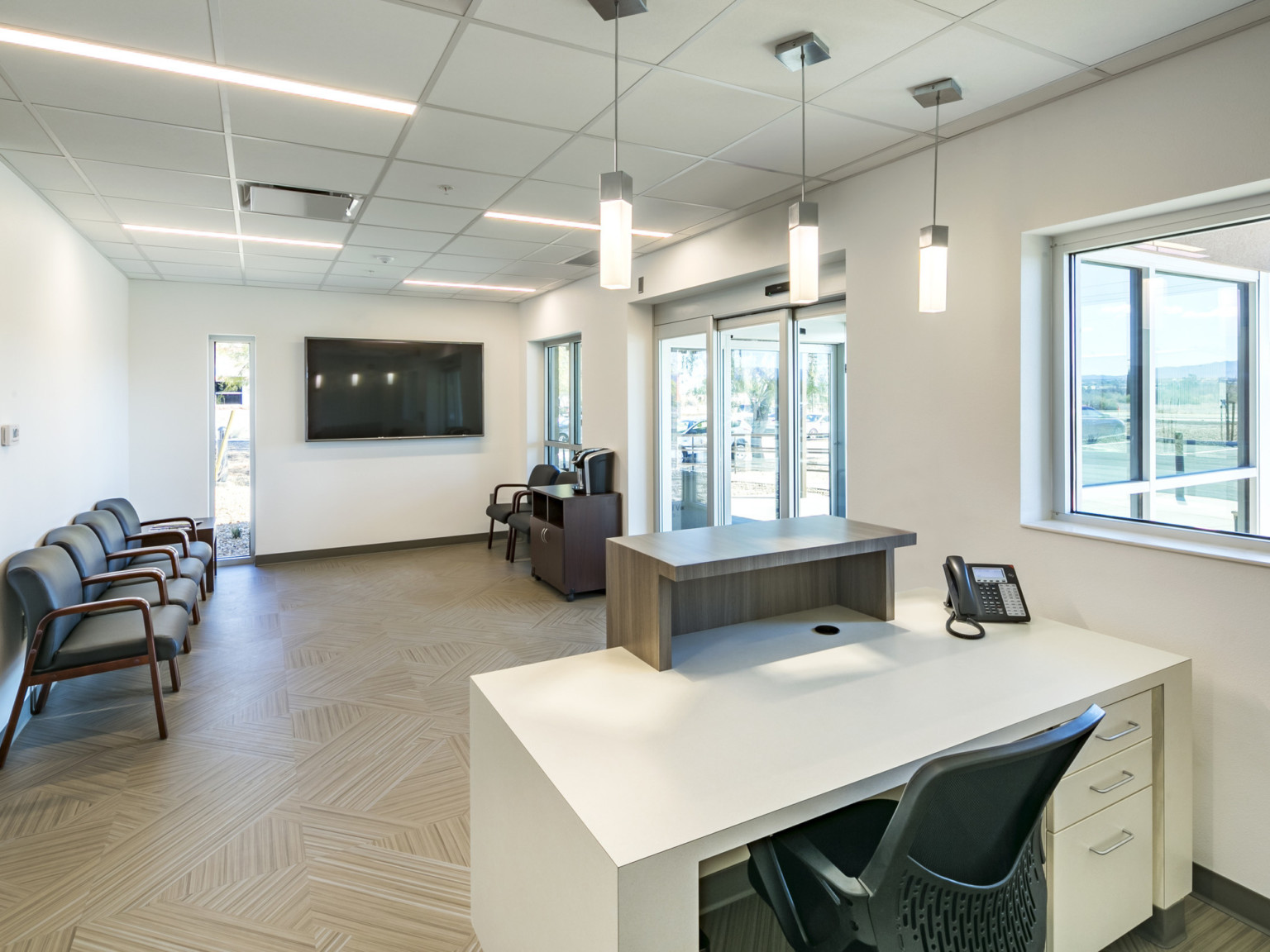 White desk with wood divider at front of white waiting room. Black chairs line left wall on wood floor facing glass doors