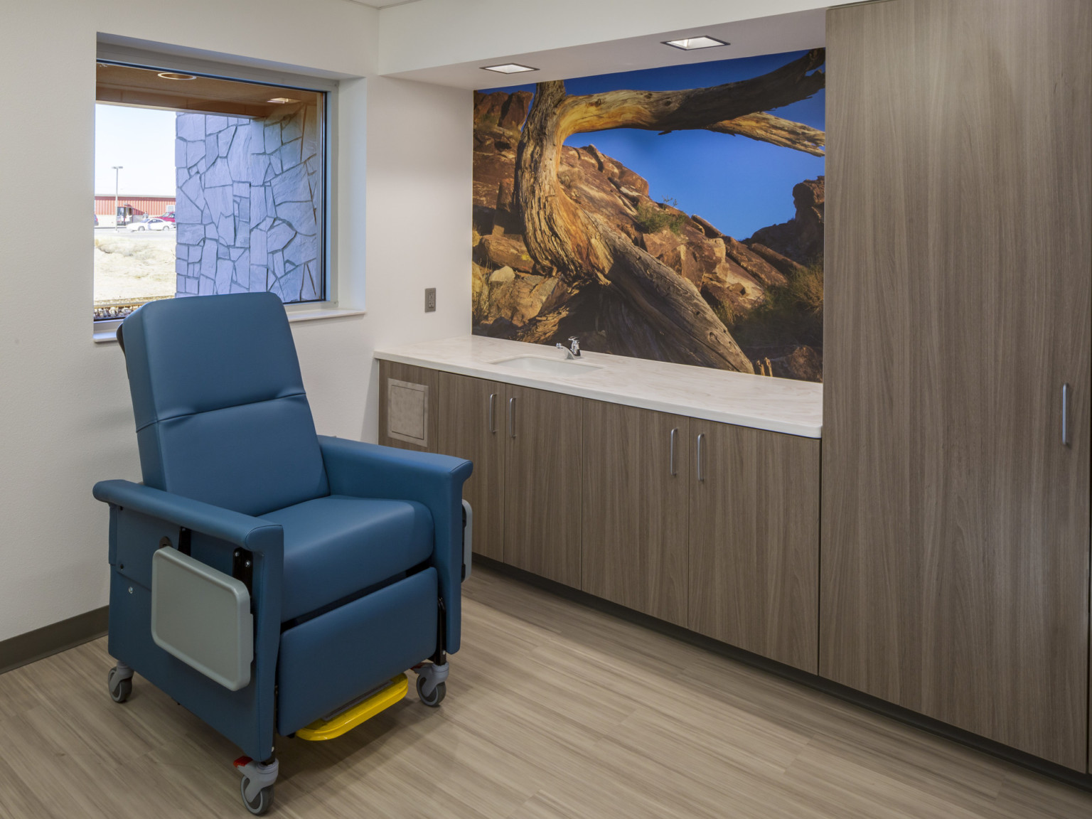 Room with blue arm chair with folded trays at side in front of window. Photo mural on wall above sink with brown cabinets