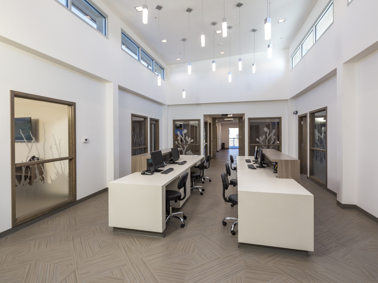 White hallway with wood details, bands of windows at the top, and hanging pendant lights. 2 long tables with chairs in center