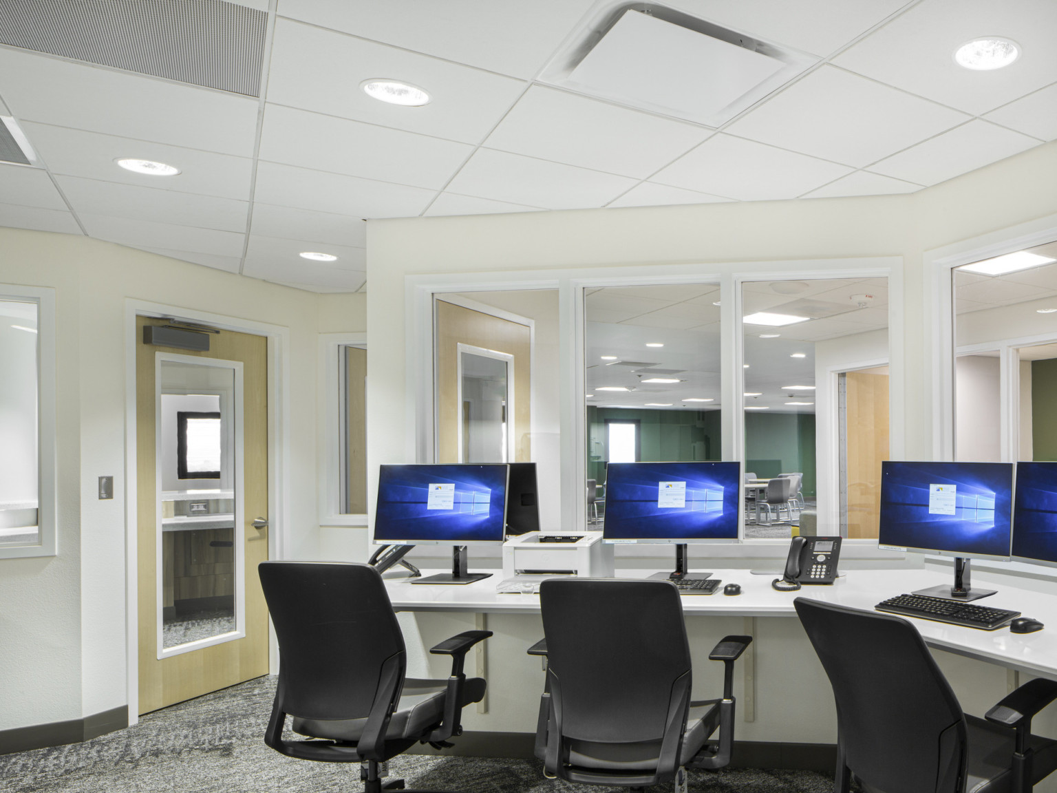 Irregularly shaped white office with desk along wall facing windows to hallway. 3 computers and chairs at desk