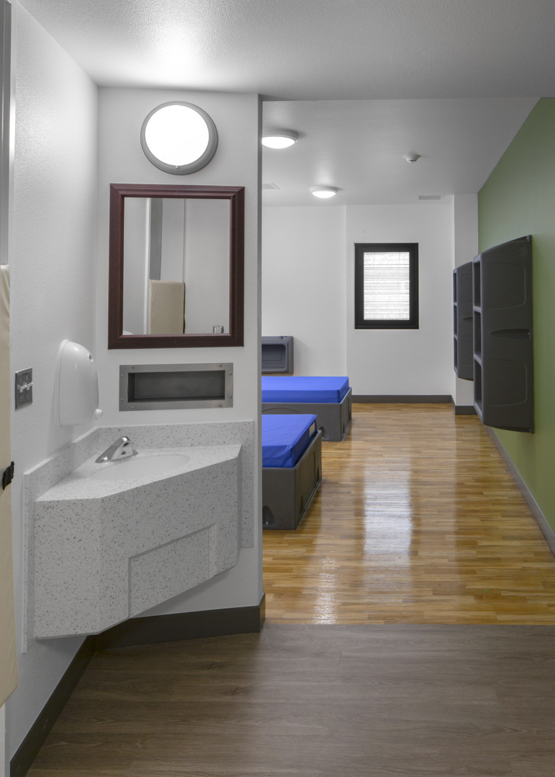 Patient room with grey sink in front, mirror and lamp on wall. Partial view of beds on grey base. Grey shelves on green wall.