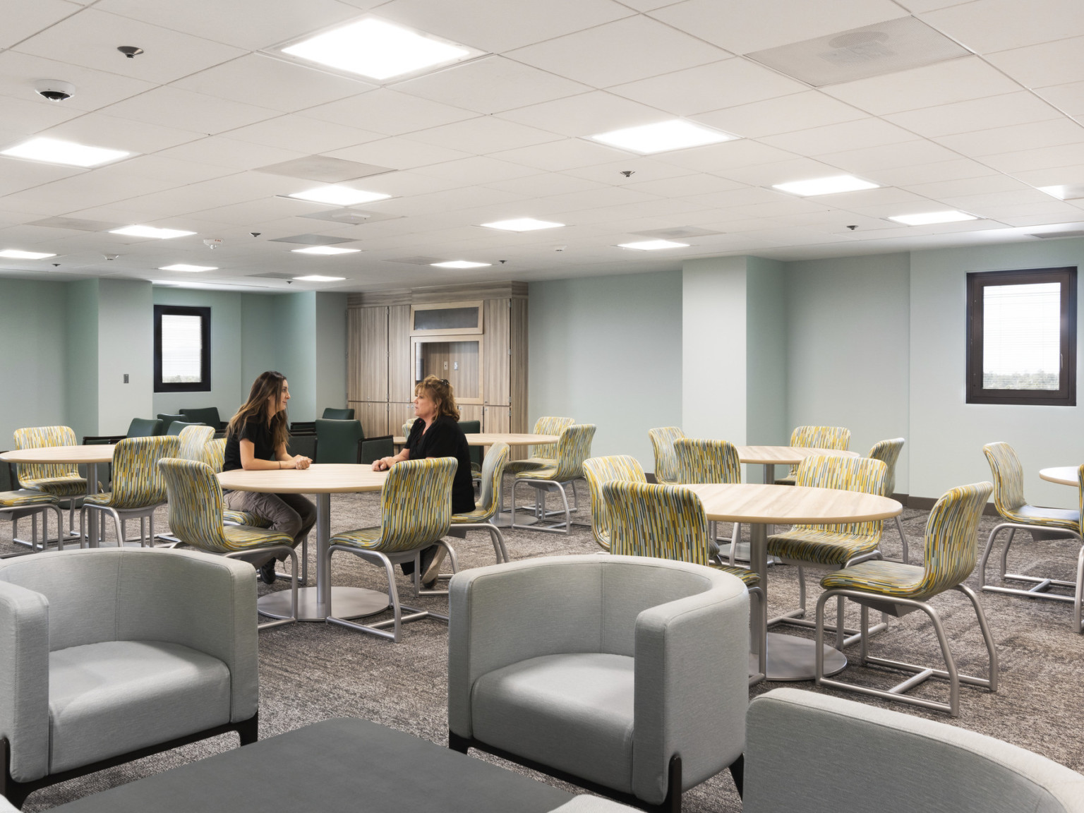 Seating area with armchairs in front, table and chairs behind. Pale blue walls with small windows. Wood cupboard in back