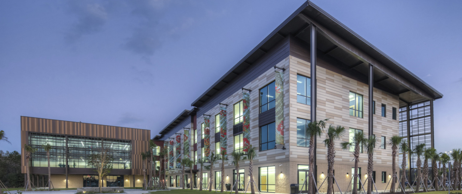 Exterior corner view of stone building lined with palm trees. Brown paneled building behind with double height windows