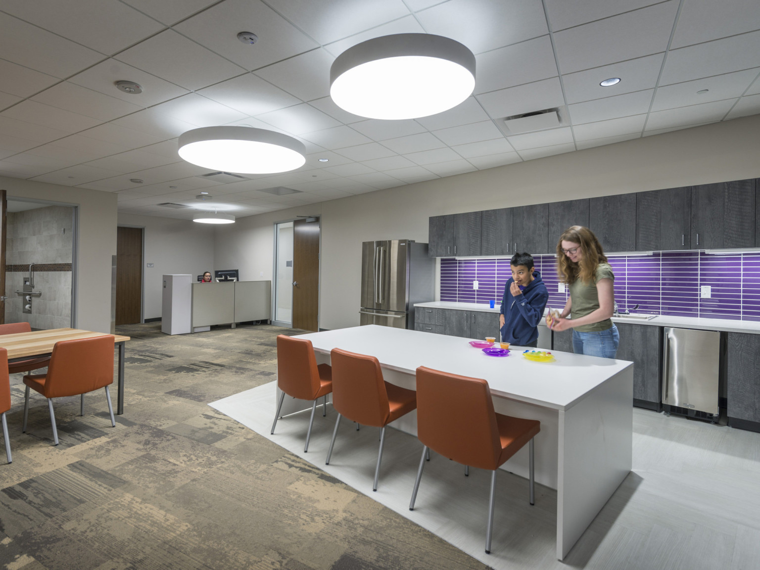 In interior office, kitchen with grey cabinets and purple backsplash. An island counter sits in front with orange chairs