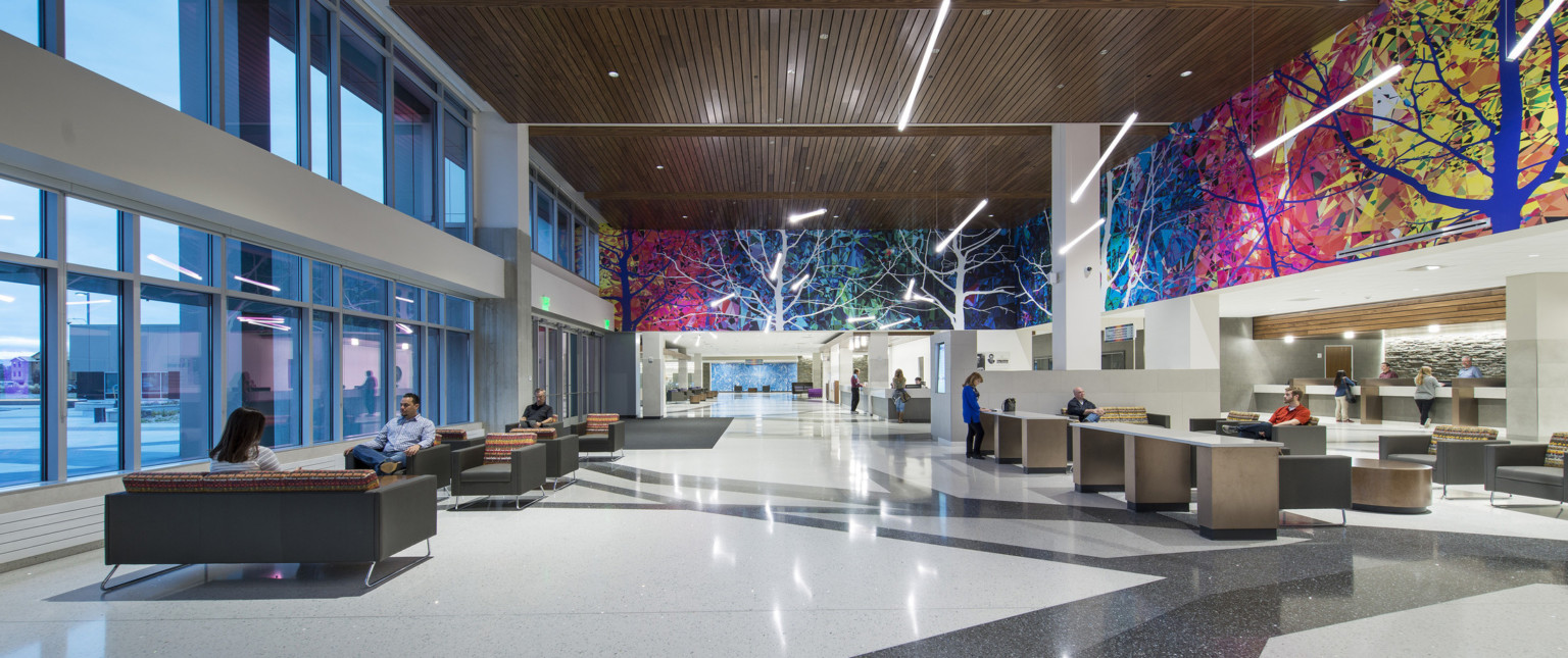 Interior lobby with colorful mural, seating space, desks, and wood detailed ceiling. Tall wall of windows lines the left wall