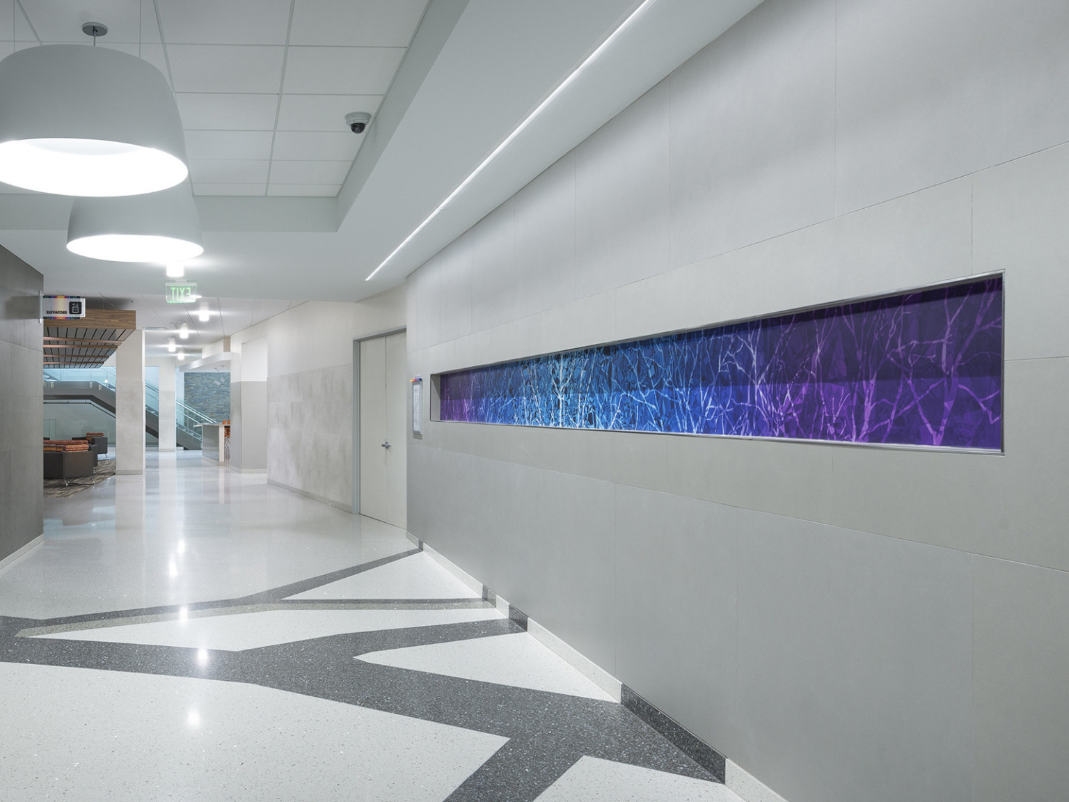 Line pattern on floor leading up to grey wall of hallway with stripe of blue and purple mural, circular light fixtures above